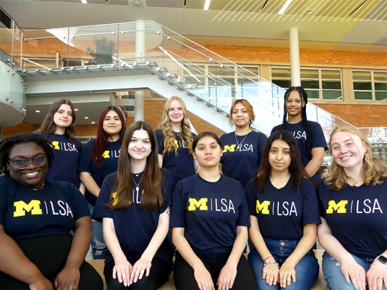 Ten female undergraduate students, wearing blue LSA t-shirts with a maize Block M, pose for a photo inside the LSA building. Five students are sitting down and five students stand behind them.