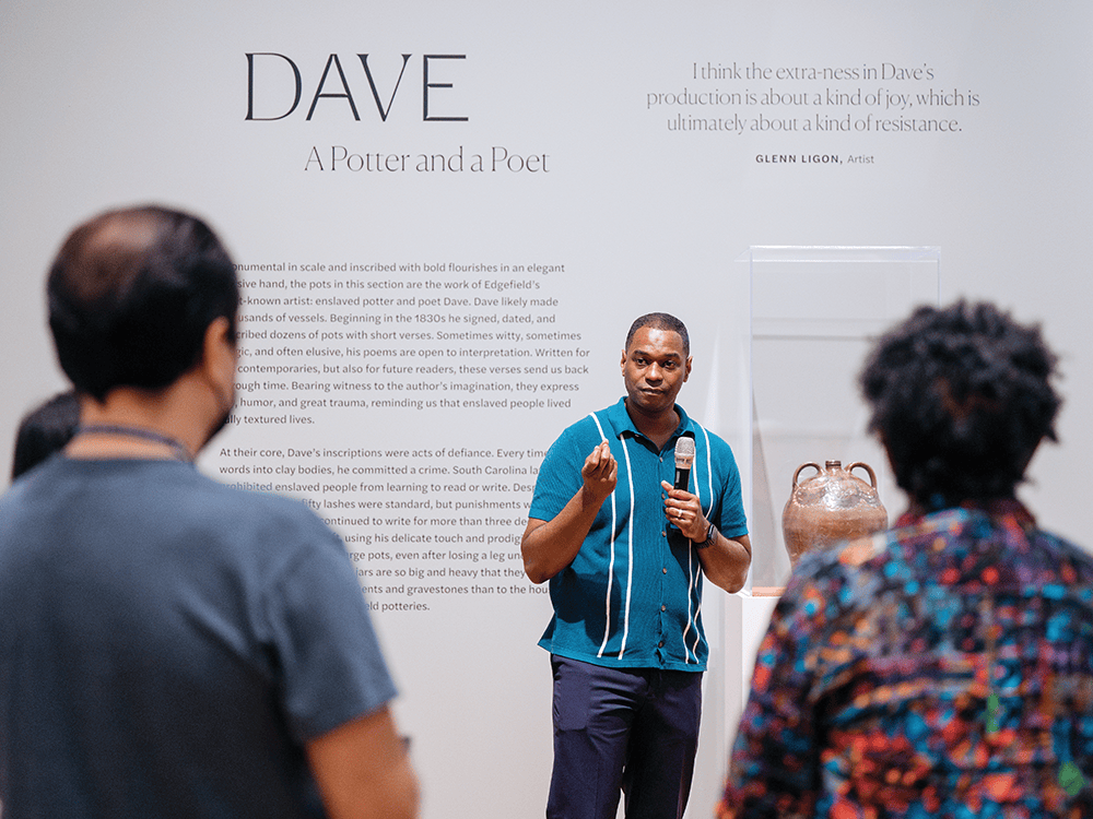 In this photo, Professor Jason Young grips a microphone with one hand while standing in front of a museum wall that reads Dave: A Potter and a Poet. With his other hand, he presses an index and middle finger against his thumb and holds his hand up as if handing over a small ring or some other prized possession. In the foreground, two visitors to the gallery look on intently.