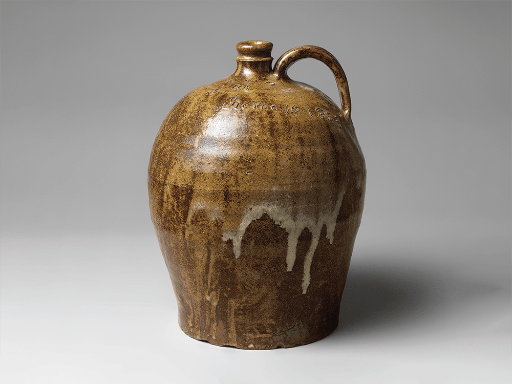 In this photo, a beige jug with a handle is mottled with darker shades of brown. Alkaline glaze drips from the pot’s center and potter Dave’s handwriting ornaments the top.