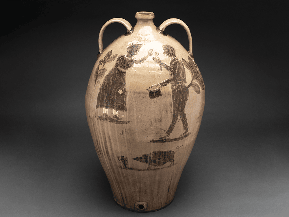 In this photo, a large watercooler jar is adorned with patterning and two figures, a man and a woman, who look to be represented as partners getting married. The two hold glasses up as if toasting one another. Beneath the two a large hog and another pot, much like those made in Edgefield, South Carolina, face each other.