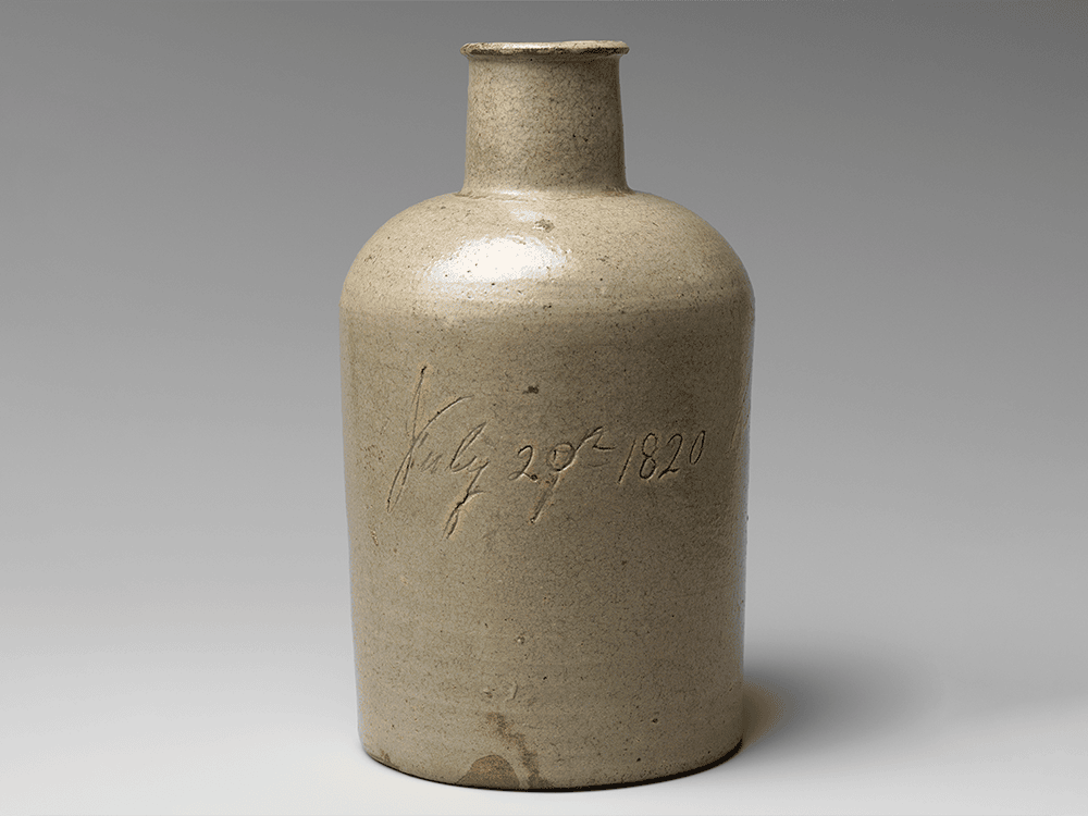 A grayish brown cylindrical and bottlelike pot shines against a stark background in this photograph. Across the front of the bottle an inscription reads, July 20th, 1820.