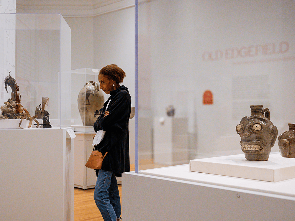 A lone woman looks at an art piece behind glass in this photo. In the foreground, face vessels are illuminated by overhead lights. The words Old Edgefield are just out of focus on a rear wall.