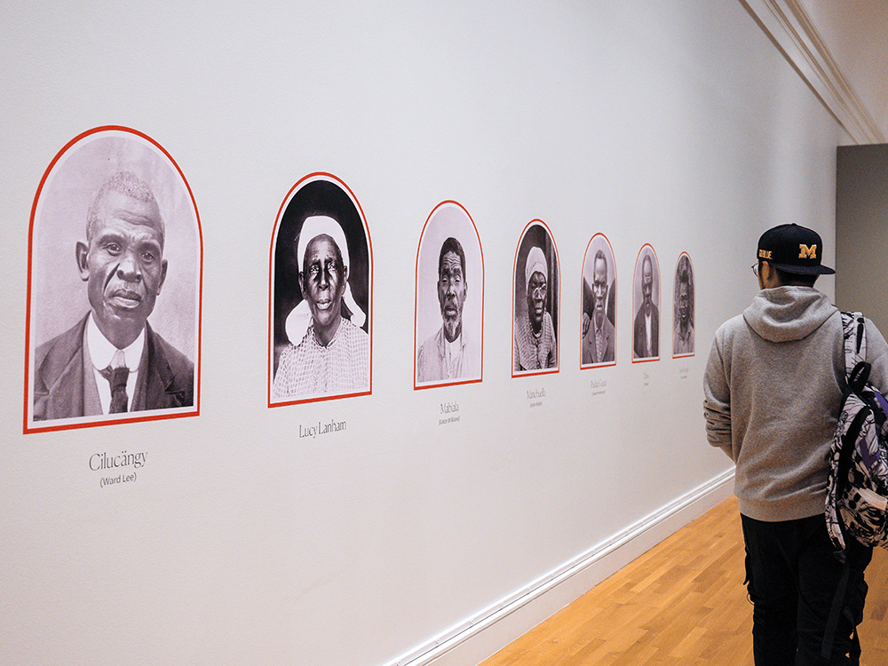 On a long museum wall, black and white photos of formerly enslaved individuals are framed in little orange archways in this photo. A man in a University of Michigan Block M cap, carrying his backpack, appears to be walking alongside the wall with the photos and reading their titles. 