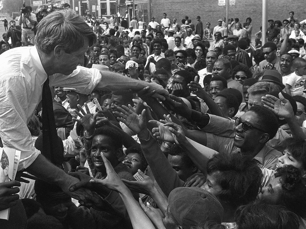 In this black-and-white photo, Presidential candidate Bobby Kennedy leans in to a crowd of hundreds of people, mostly people of color, in Detroit, reaching forward to shake hands during a campaign stop. He is wearing shirt sleeves and sports a slight smile.
