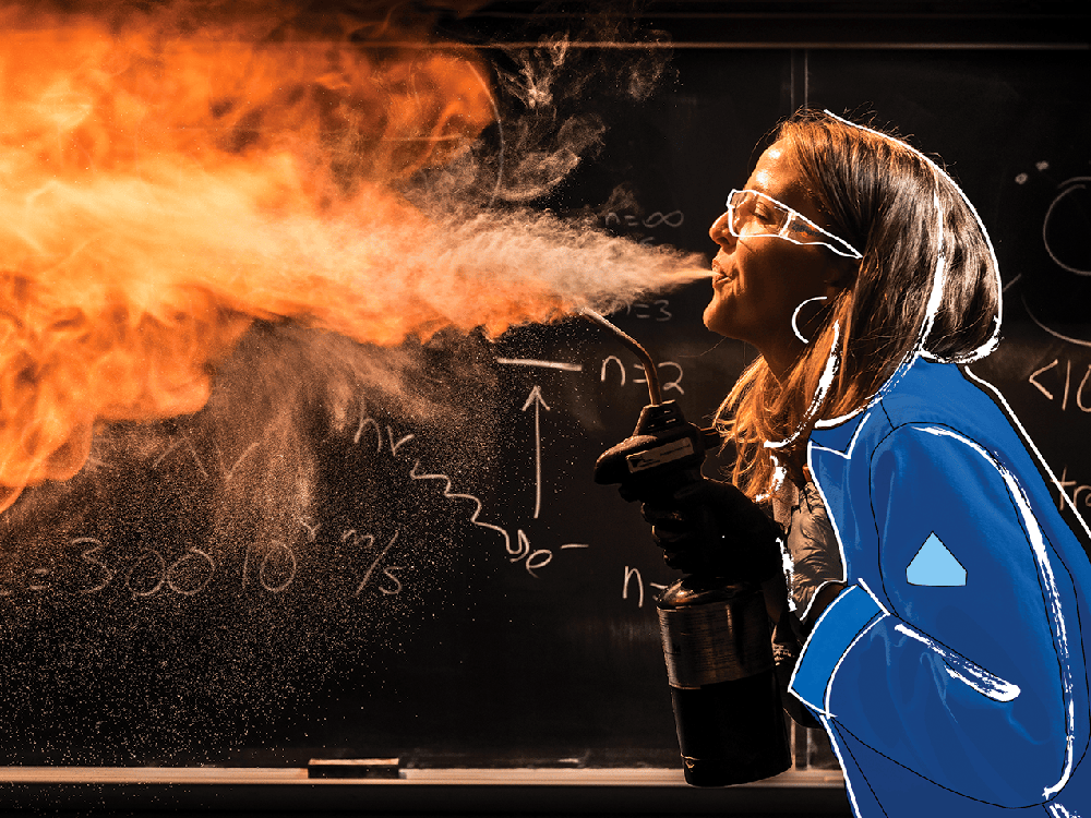 In this photograph, Kate Biberdorf wears a dark blue fire safety suit and protection goggles as she stands in front of a black chalkboard with white writing all over it. She faces to the left, holding a blowtorch angled upward toward her mouth, appearing to breathe out fire across the page.