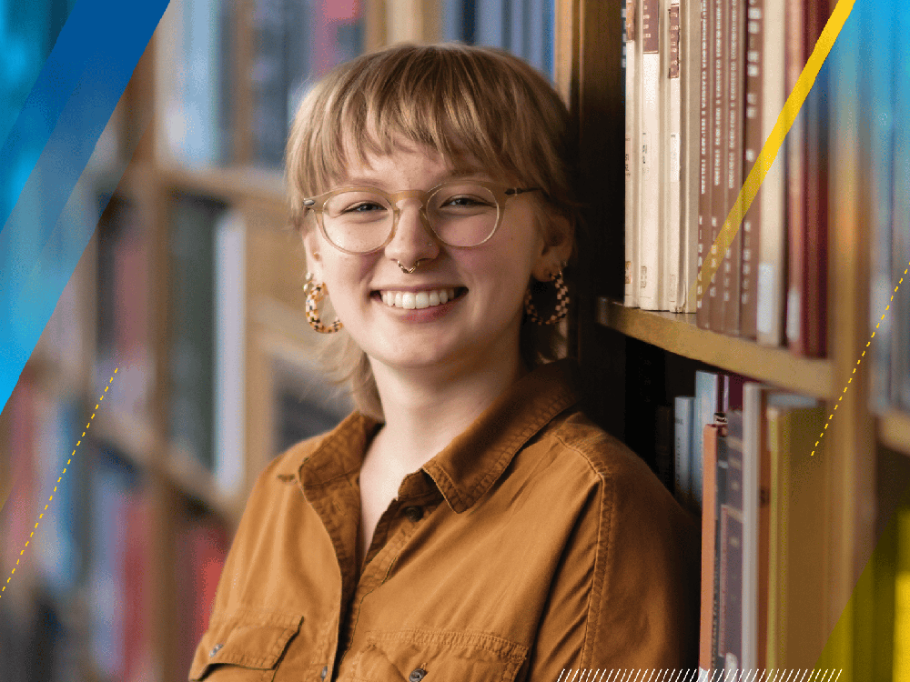 A photograph of Cassidy, a U-M undergraduate student, who has short blond hair and wears glasses. They are wearing a short-sleeved copper-colored shirt. They are smiling in the photo.