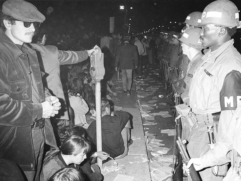 A black-and-white photograph shows a tense standoff between a protester and military police at the 1968 Democratic National Convention. It is dark outside. The white protester wears dark glasses, a cap, and a corduroy jacket. Other protesters sit and stand near him. The stoic military police wear uniforms that say “U.S. Army.” The one who is central to the photo is a man of color. Papers are strewn on the sidewalk and street.