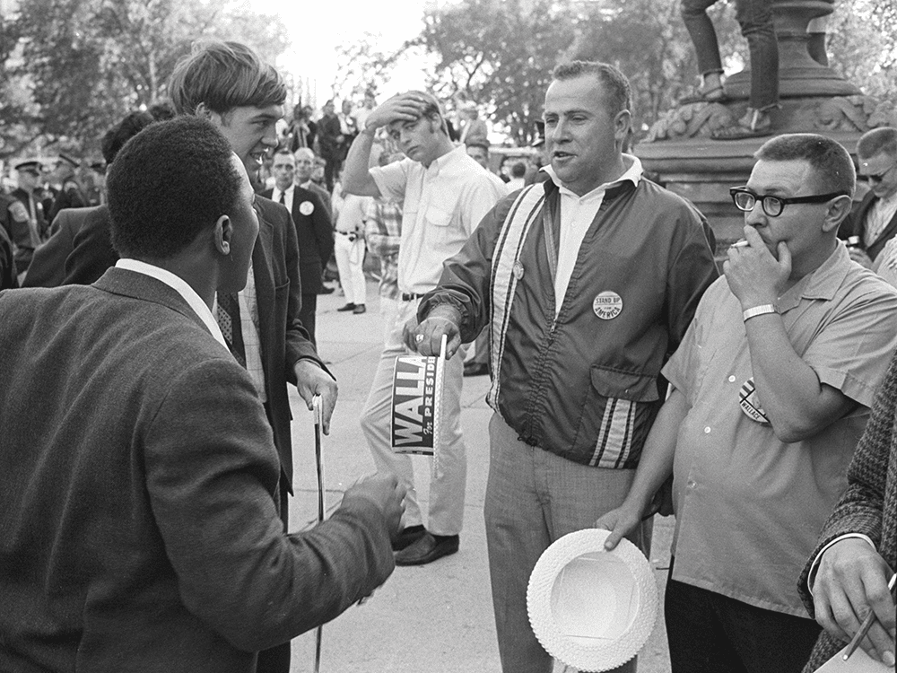 In this black-and-white photo, two men—a man of color and a white man—face two white men who hold “Wallace for President” hats. They appear to be arguing. The image was made at a campaign event for George Wallace in Lansing, Michigan.