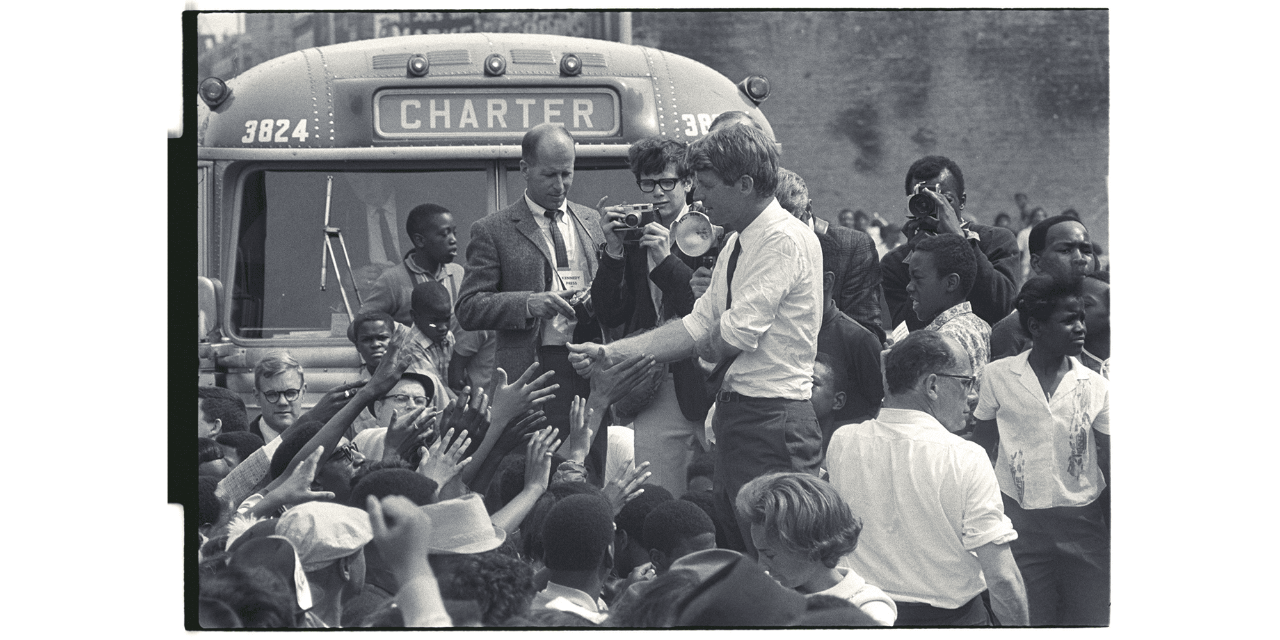 In this black-and-white photo, Presidential candidate Bobby Kennedy stands elevated amid a diverse crowd, with many people reaching up to shake his hand. Photographer Jay Cassidy, a student at the University of Michigan, faces the camera while holding a camera of his own. Another photographer stands next to Cassidy. Behind them is a bus that says “Charter.” 