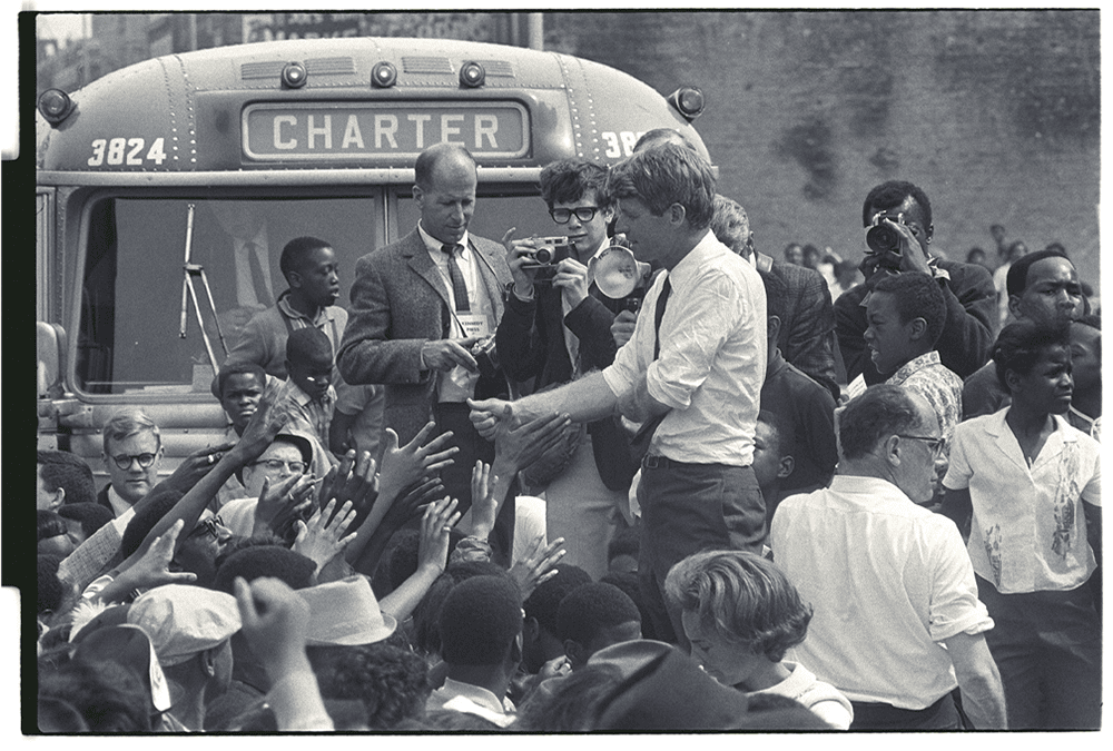 In this black-and-white photo, presidential candidate Bobby Kennedy stands elevated amid a diverse crowd, with many people reaching up to shake his hand. Photographer Jay Cassidy, a student at the University of Michigan, faces the camera while holding a camera of his own. Another photographer stands next to Cassidy. Behind them is a bus that says “Charter.”