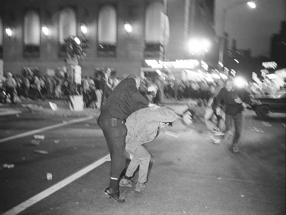 In this black-and-white photo, a police officer overpowers a protester in the street at the 1968 Democratic National Convention in Chicago. It is dark outside, and the two are standing in the street. Around them are blurry images of people scattering under the streetlights.