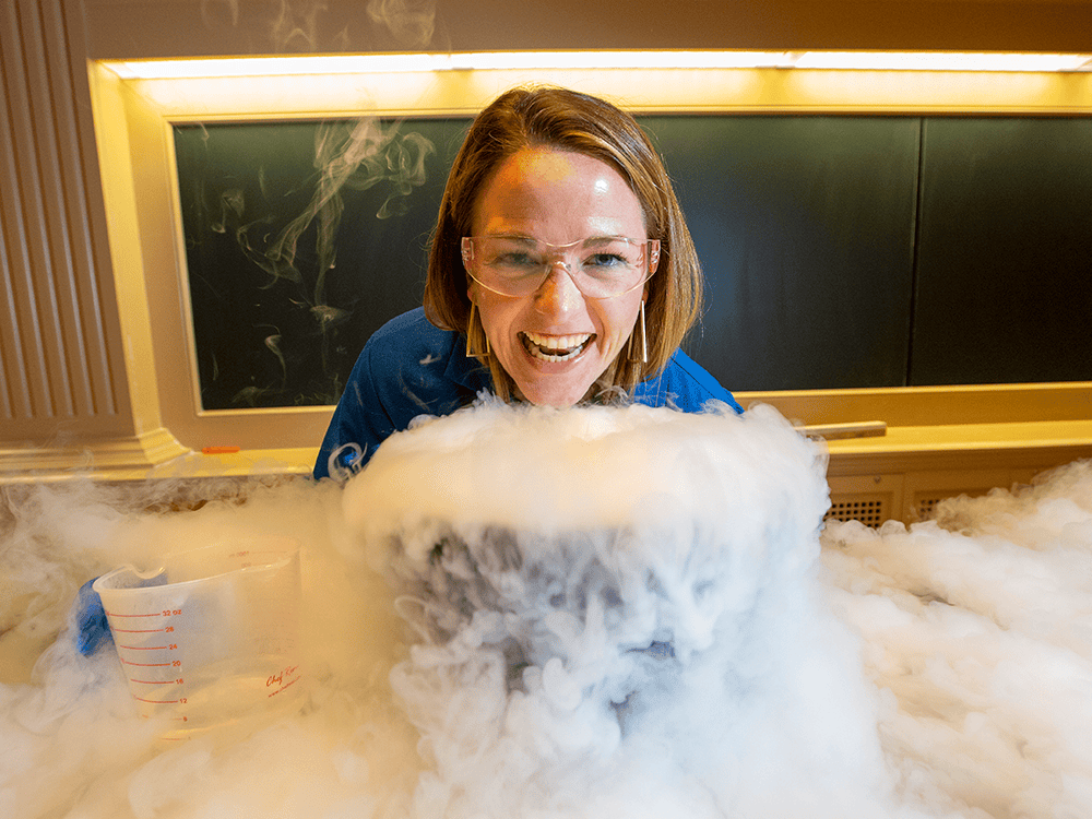In this photograph, Kate Biberdorf wears a dark blue safety suit and safety goggles and smiles as she stands behind a large bucket on a classroom desk that spews liquid nitrogen all around her. The professor is in a classroom in front of a clean chalkboard.
