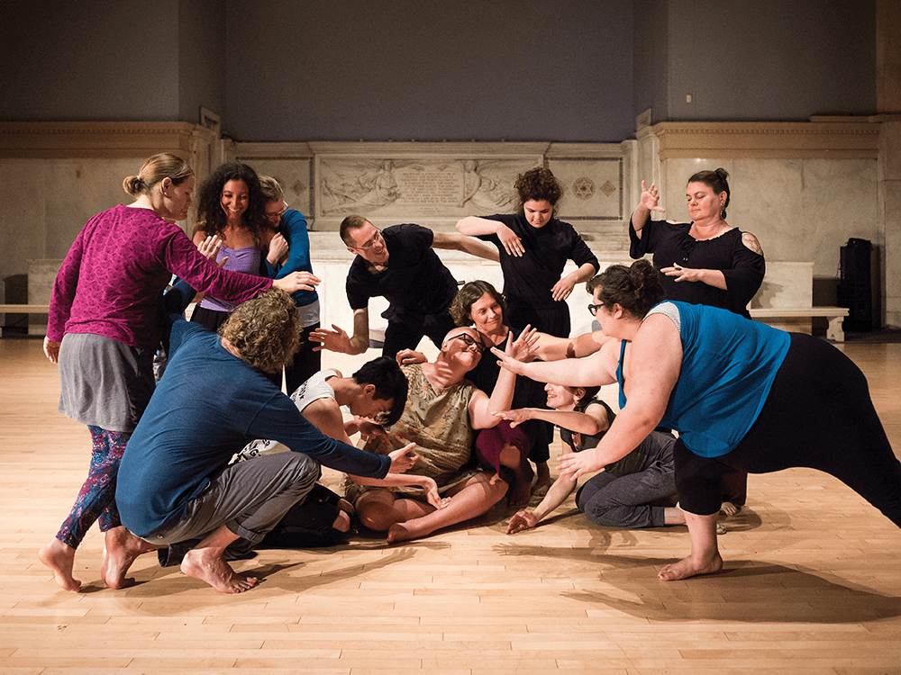 A photo shows a group of people moving together during an Asylum Project dance at Judson Church. The group moves atop a wooden floor, with marble walls in the background. Some are sitting, others standing, but all face Professor Petra Kuppers and her wife, who are at the center. The diverse group’s swirl patterns, arm linking, and eye connections are prevalent, forming something akin to the form of a flower.