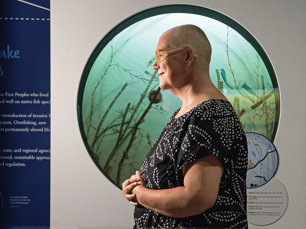  For this photo, Professor Petra Kuppers stands in front of a circular aquarium window in the U-M Museum of Natural History. The light hits her face from one side of the room and her profile is illuminated from behind where water-behind-glass catches light from the surface above. On the blue and white wall are captions about the state of Michigan’s land and history.