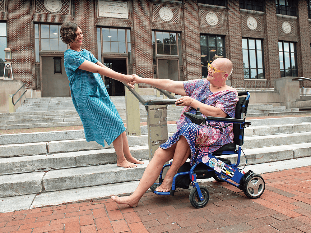 In this photo, Professor Petra Kuppers and her wife hold hands and lean outward from one another. Petra sits in her blue mobility scooter and her wife stands on stairs that lead to Hatcher Graduate Library’s entrance. They hold each other’s gaze and smile at one another.