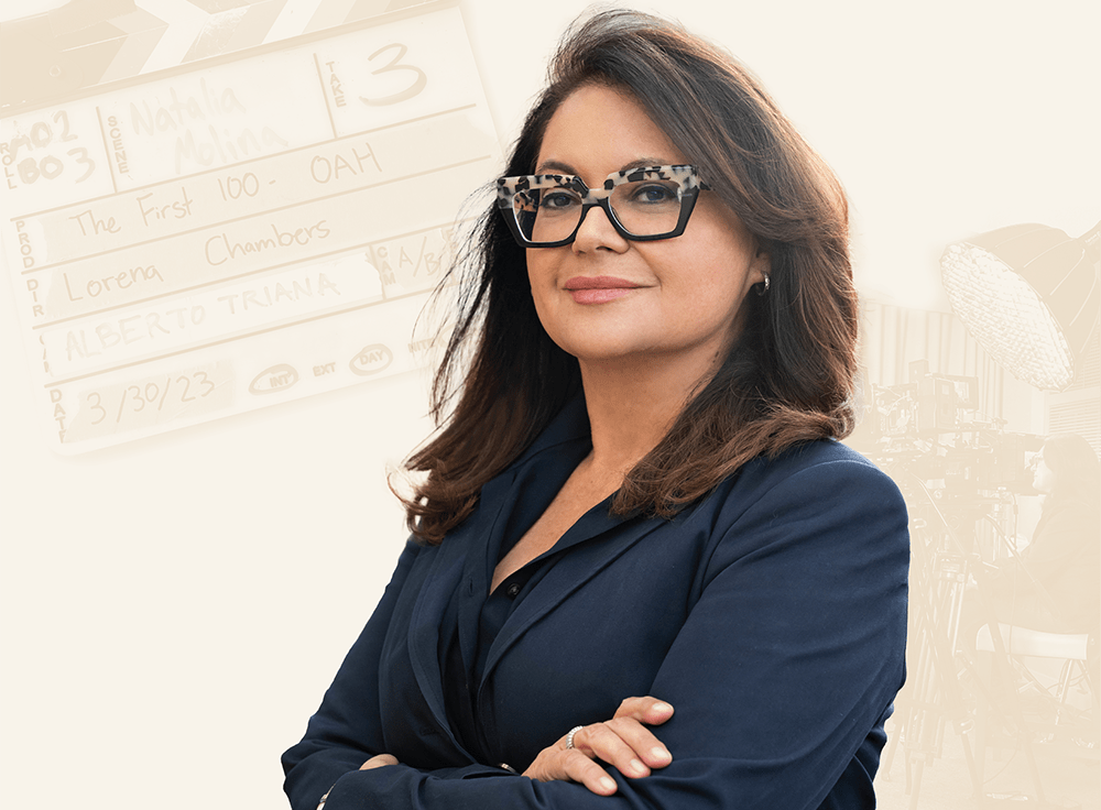 In a photograph, Lorena Chambers stands with her arms crossed in a blue blazer. She also wears a leopard patterned and black-rimmed pair of glasses and gazes at the camera. In the background is a movie clapperboard. The clapperboard has readouts about the production of Chambers’s project The First 100. In the background, a film crew with lighting and cameras works within a studio production space.
