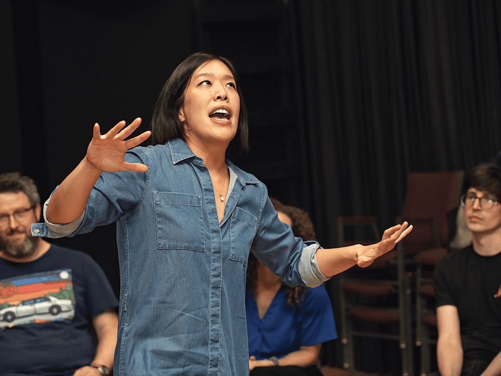 Lillian Li, a woman with dark hair, wears a denim shirt and stands on a stage, gesturing with her arms.