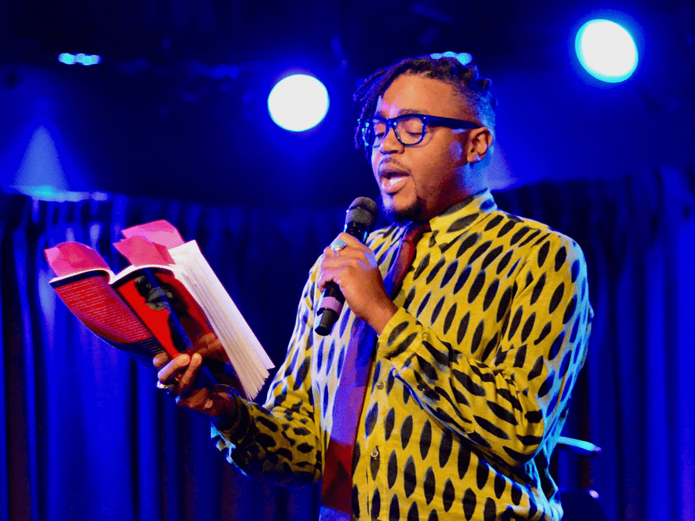 Darrel Alejandro Holnes stands on a blue-lighted stage, wearing a yellow and black shirt, and reading into a microphone.