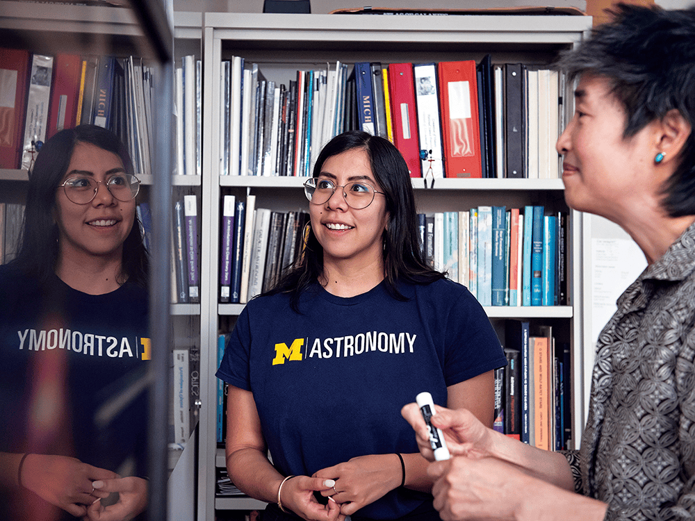 In front of a bookcase in Professor Sally Oey’s office, astronomy Ph.D. student Irene Vargas-Salazar, who has long, straight black hair and glasses, looks at a white board with Oey toward the left side of the image. Both Vargas-Salazar and Oey are smiling. Oey is holding a black EXPO marker and Vargas-Salazar is wearing a U-M Astronomy shirt.