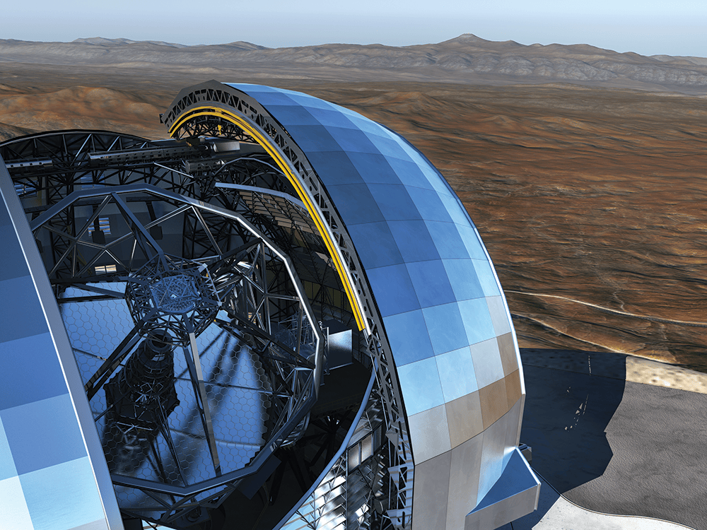 The top of the Extremely Large Telescope faces the camera, angled toward the sky and upper right corner of the image. Two large, spherical mirrors sit on each side of the top of the telescope. Behind the telescope is a landscape of the Chilean Atacama Desert.