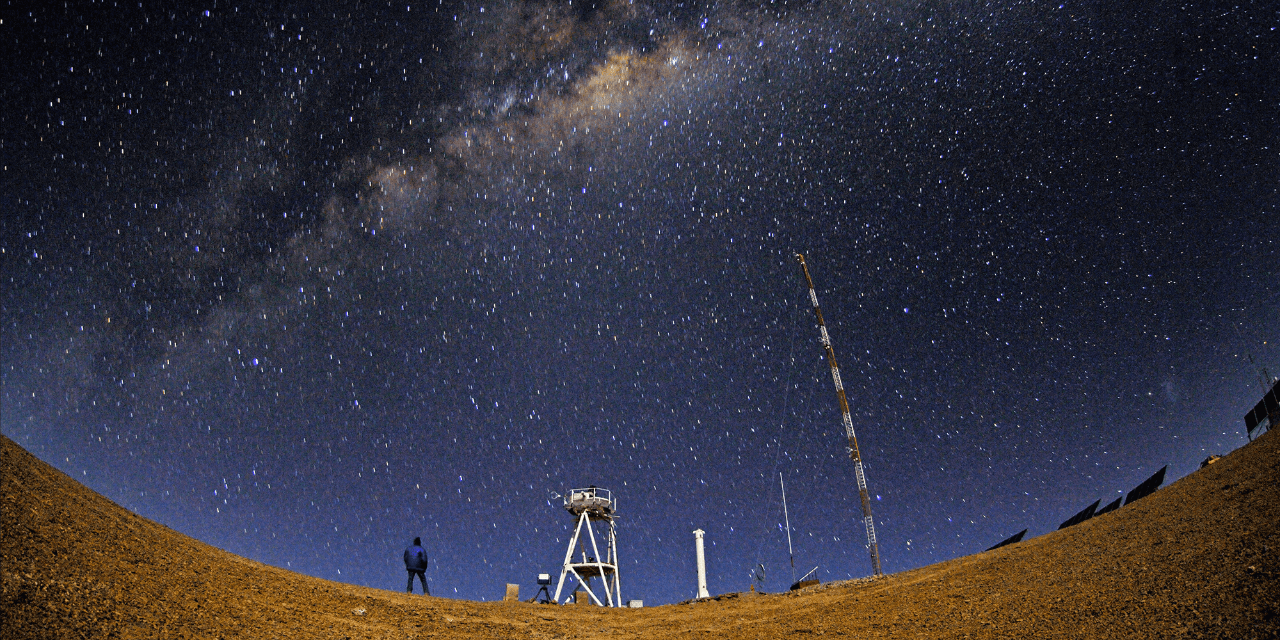 A nighttime panoramic view of the Chilean Atacama Desert with small stars scattered across the sky. A person in a dark blue jacket and dark pants looks up at the sky where a clear band of the Milky Way galaxy is in the middle of the image. In front of the person in the middle ground is a white watchtower and other equipment.