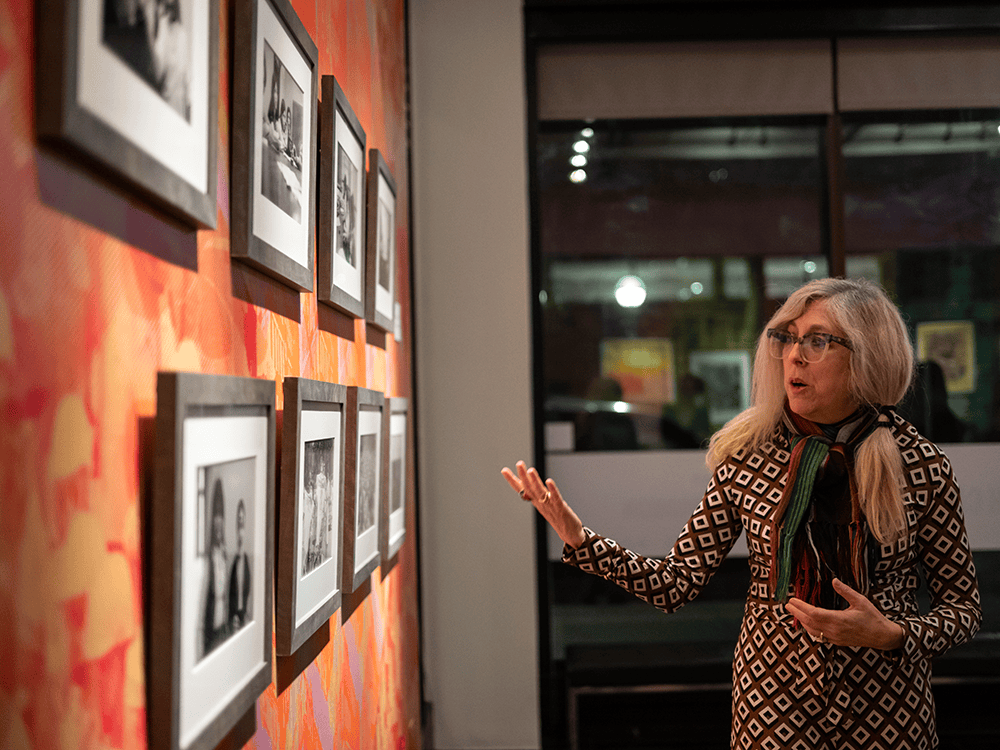 Amanda Krugliak, arts curator and assistant director of arts programming at the Institute for the Humanities, stands next to photographs on a wall. Krugliak wears glasses, a patterned jumpsuit, and a scarf. The photos are black and white, and the wall is orange. 