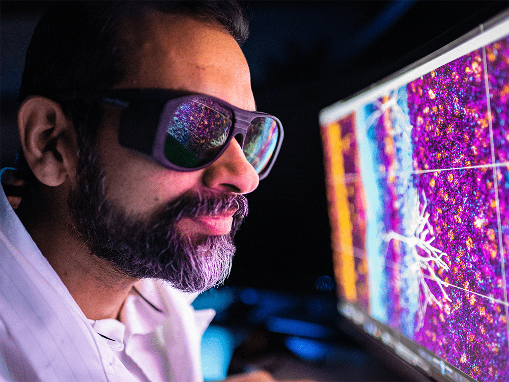 An environmental portrait shows Professor Omar Ahmed in profile, wearing dark glasses that reflect the images on a monitor he is viewing. The image shows the retrosplenial cortex in vibrant shades of purple, pink, blue, and yellow.