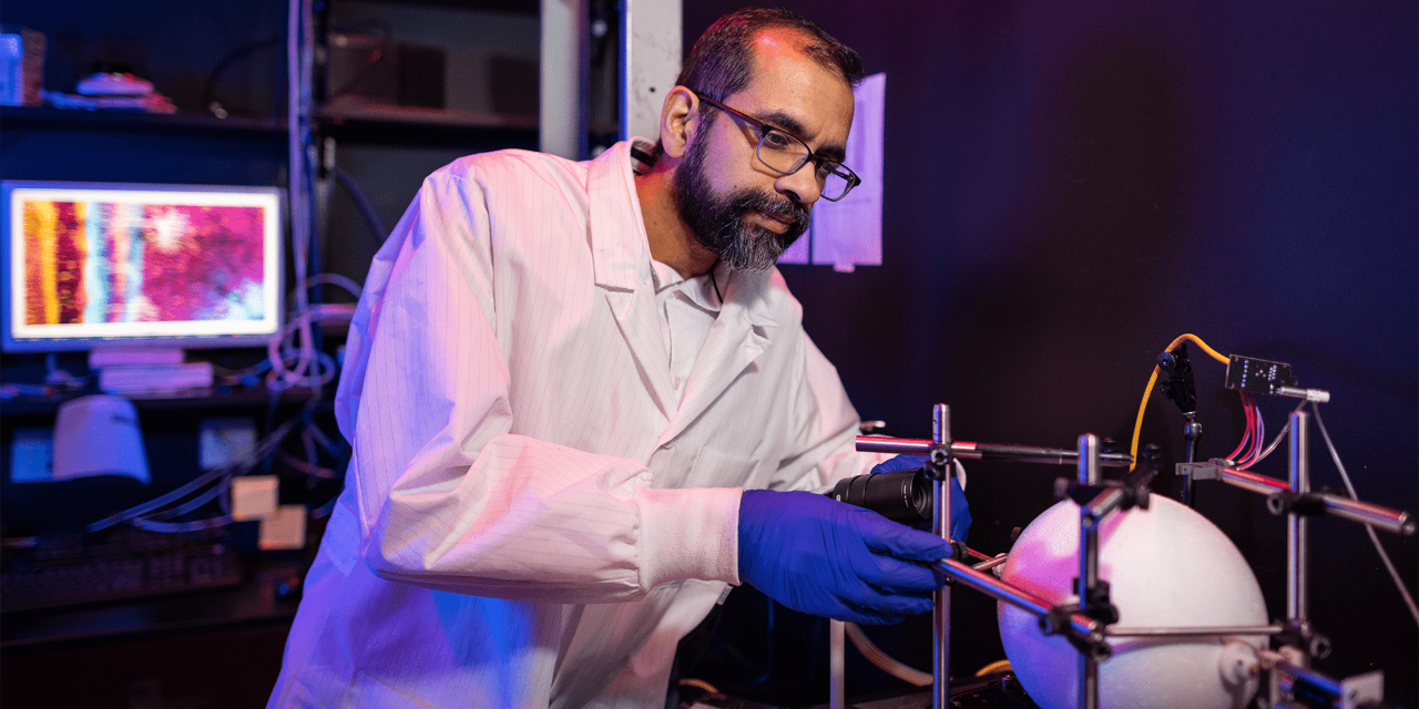 In purple lighting, Professor Omar Ahmed adjusts a lab instrument for testing brain activity. He is wearing a lab coat and blue gloves. A computer monitor is casting light out into the darkness behind him. Various pieces of lab equipment and other items are stacked on shelves that are attached to the rear wall of the room.