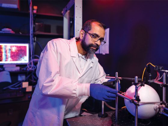 In purple lighting, Professor Omar Ahmed adjusts a lab instrument for testing brain activity. He is wearing a lab coat and blue gloves. A computer monitor is casting light out into the darkness behind him. Various pieces of lab equipment and other items are stacked on shelves that are attached to the rear wall of the room. 