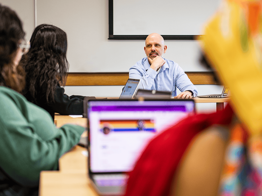 Matthew Sullivan sits at the front of the classroom and is engaged in conversation with one of his students. Others in the room look on. One of the students’ laptop screens is in view.