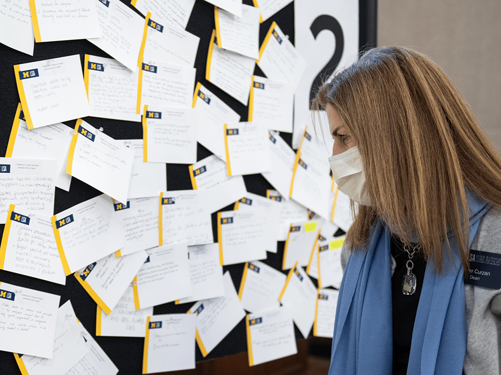 LSA Dean Anne Curzan looks at a board covered with sticky notes that include suggestions from faculty, staff, and students about how the college can improve its diversity, equity, and inclusion efforts. Dean Curzan wears a mask and is pictured in profile.