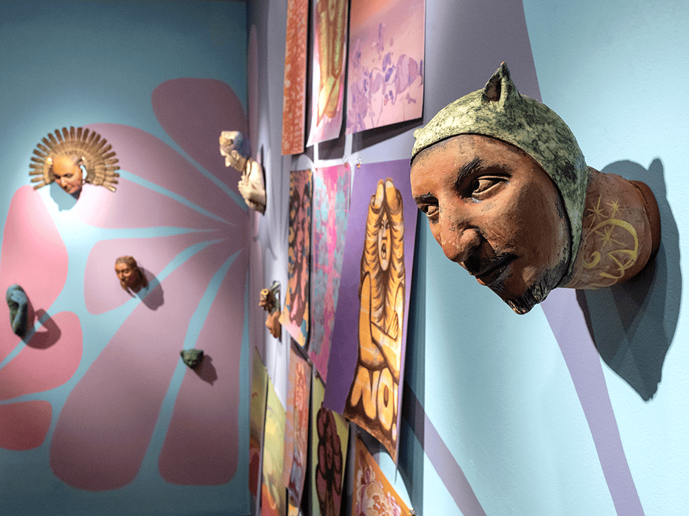 Brightly colored artwork is displayed on a wall that is light blue, pink, and purple. In the foreground is a three-dimensional ceramic piece that looks like a human male’s head coming out of the wall. In the background are paintings and more sculptural pieces. They are part of the exhibition With Care by artist Nicole Marroquin. 