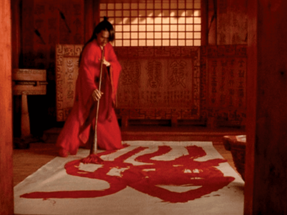 A character in the movie “Hero” wears a long red robe and paints the character character “jian,” which means “sword,” in red paint on the floor. The character is surrounded by walls that glow with red light.