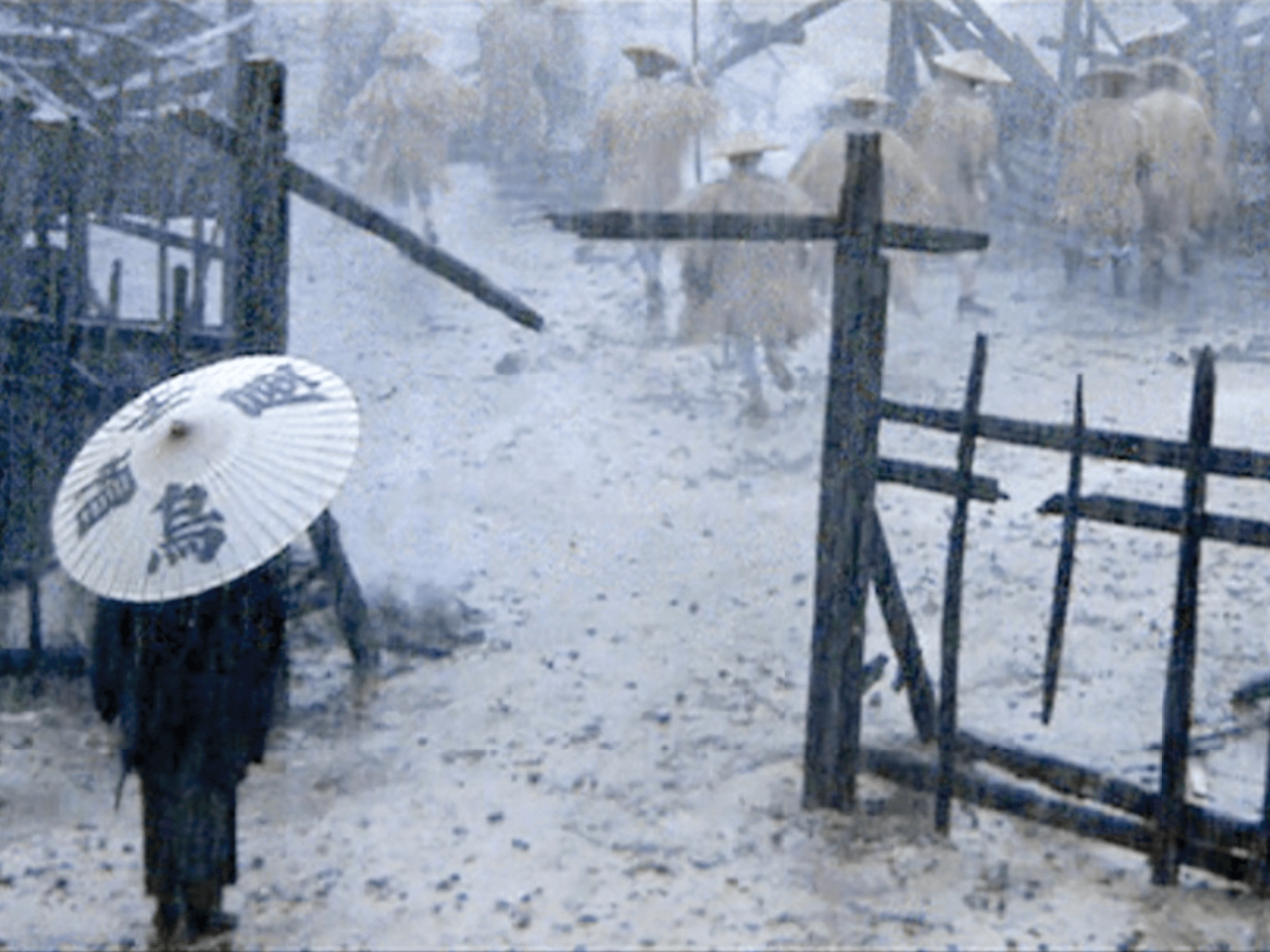 In a film still, a figure holds a white umbrella that is painted with calligraphic characters in black ink. They stand in the snow, in front of a broken fence. On the other side of the fence are several human figures dressed identically in dull yellow clothing and hats. 