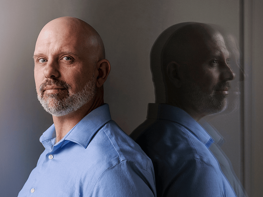Sociology faculty member Matthew Sullivan poses in a light blue dress shirt, with his reflection visible to the right of his head. His expression is serious.