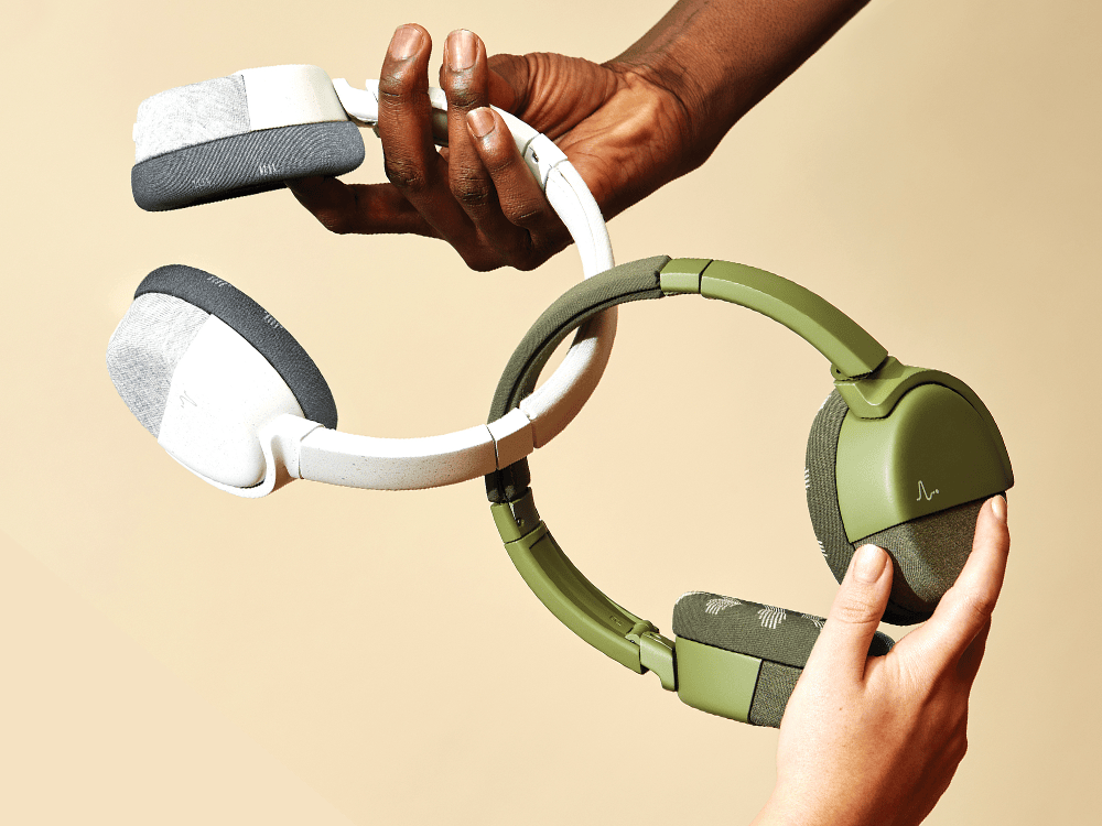 Two pairs of wireless over-ear headphones are intertwined, the left one white and the right one green. The white headphones are held by a Black hand coming from the top of the image and the green headphones are held by a white hand coming from the bottom of the image.