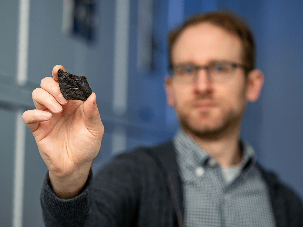 Facing the camera, Professor Matt Friedman holds a small, black fossil between his index and pointer finger. The fossil looks like an arrowhead with smoother edges. Friedman’s hand and the fossil are in focus while Friedman’s body and face are out of focus.