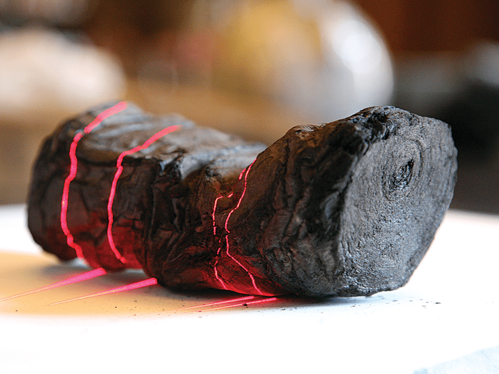 A dark brown ancient scroll is shown being scanned, with pink laser lines pointed at portions of the scroll. 