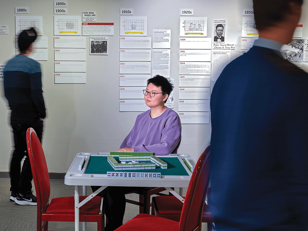 Lily Jiale Chen sits at a mahjong table in the “Detroit’s Chinatowns” exhibit gallery space at the Detroit Historical Museum while two blurred figures look at the exhibit.