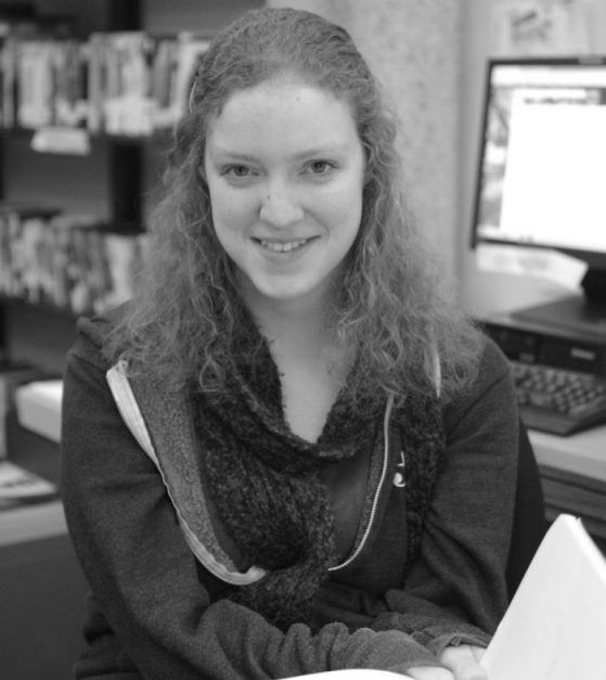 In a black and white image, Megan sits at a desk with a computer monitor in the background.
