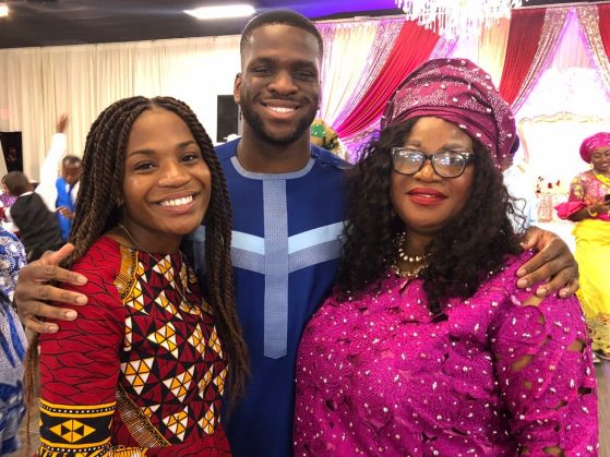 Miracle, her brother, and her mother are pictured dressed in traditional and very colorful Nigerian clothing. Miracle wears a dress of red, yellow and black geometric patterns. Her mother wears a fuchsia dress and headscarf. Her brother wears a robe in royal blue, with white and lighter blue accents. They are at a party in an opulently decorated room, with a chandelier and red, purple and gold curtains, where they are surrounded by other revelers who are wearing clothing that is equally colorful - bright yellows, pinks, cobalt blue.