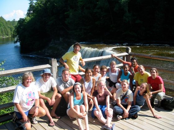 Cathy Bach is pictured with a group of UMBS students. They are posing on a wooden platform that overlooks a waterfall. It is sunny and most are wearing shorts.