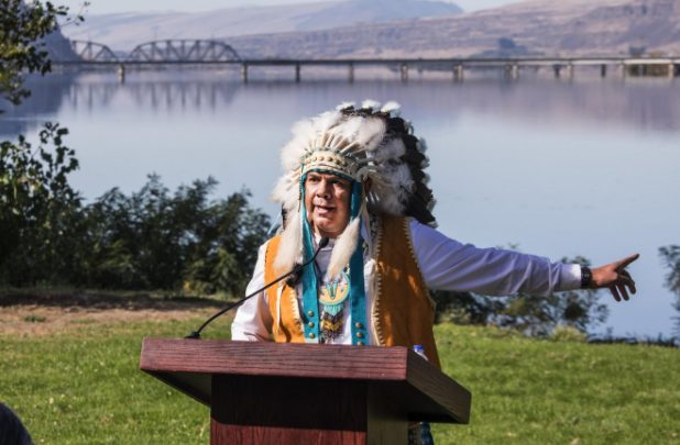 An older man in a ceremonial head dress stands at a podium gesturing to a body of water behind him
