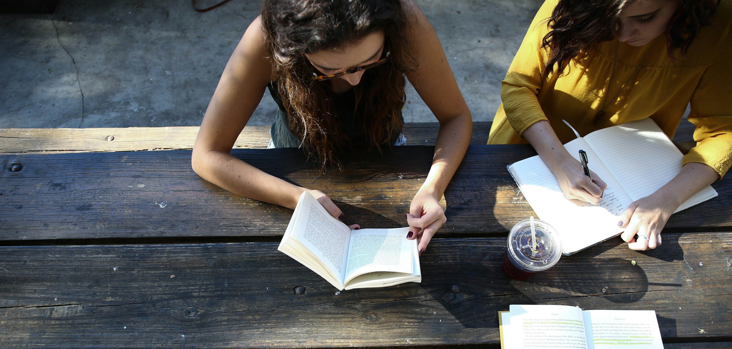 Two middle school–age children sit at a picnic table. One reads from a book, while another writes in a notebook.