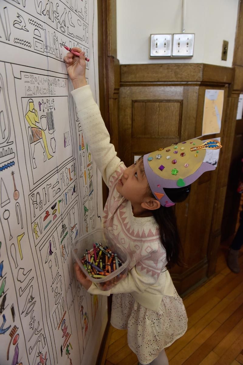 Child wearing a paper crown reaches up to color in a symbol on a large hieroglyphics coloring page.