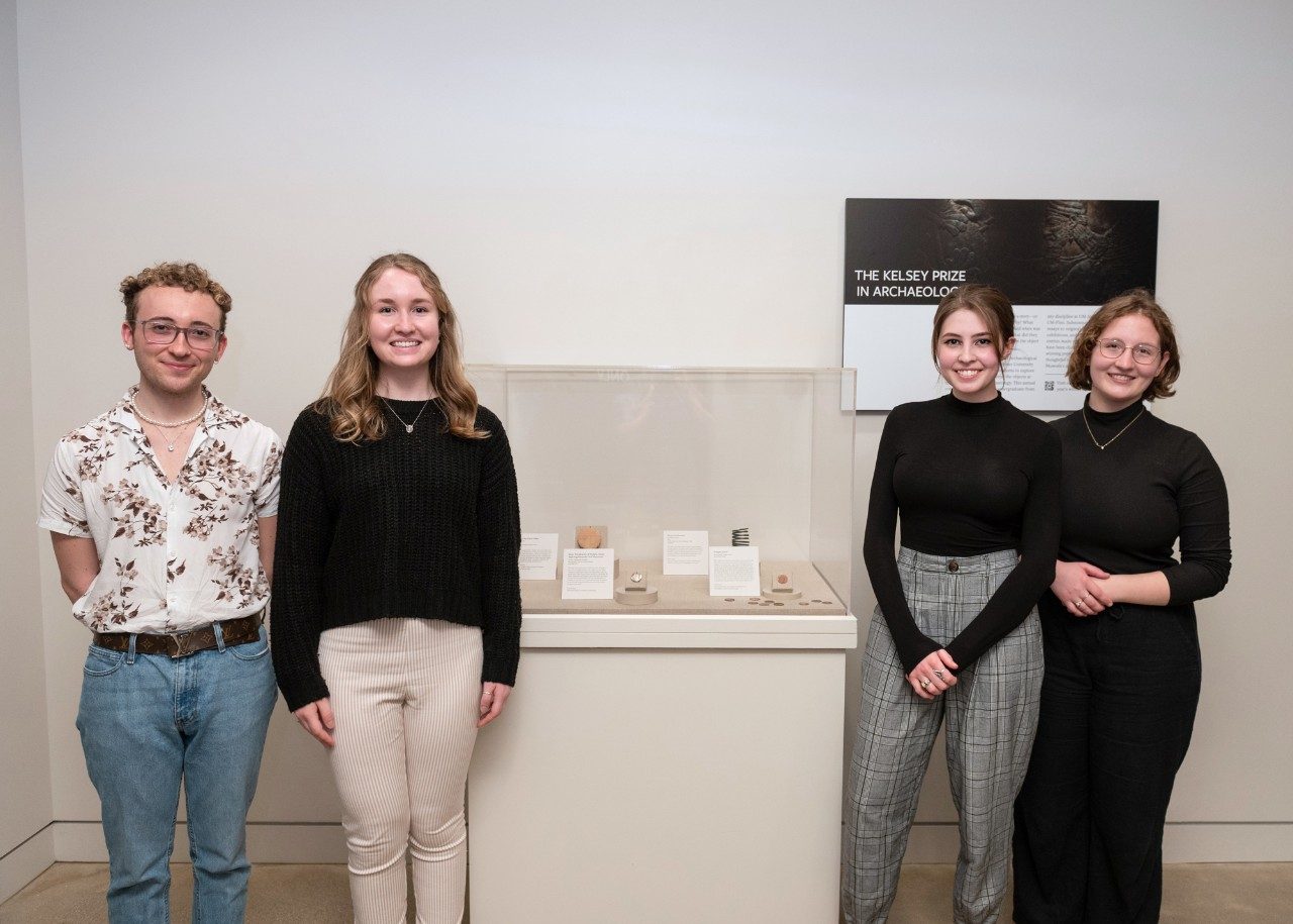 Four college-age students stand beside a small museum vitrine in filled with coins, jewelry, and a tablet.