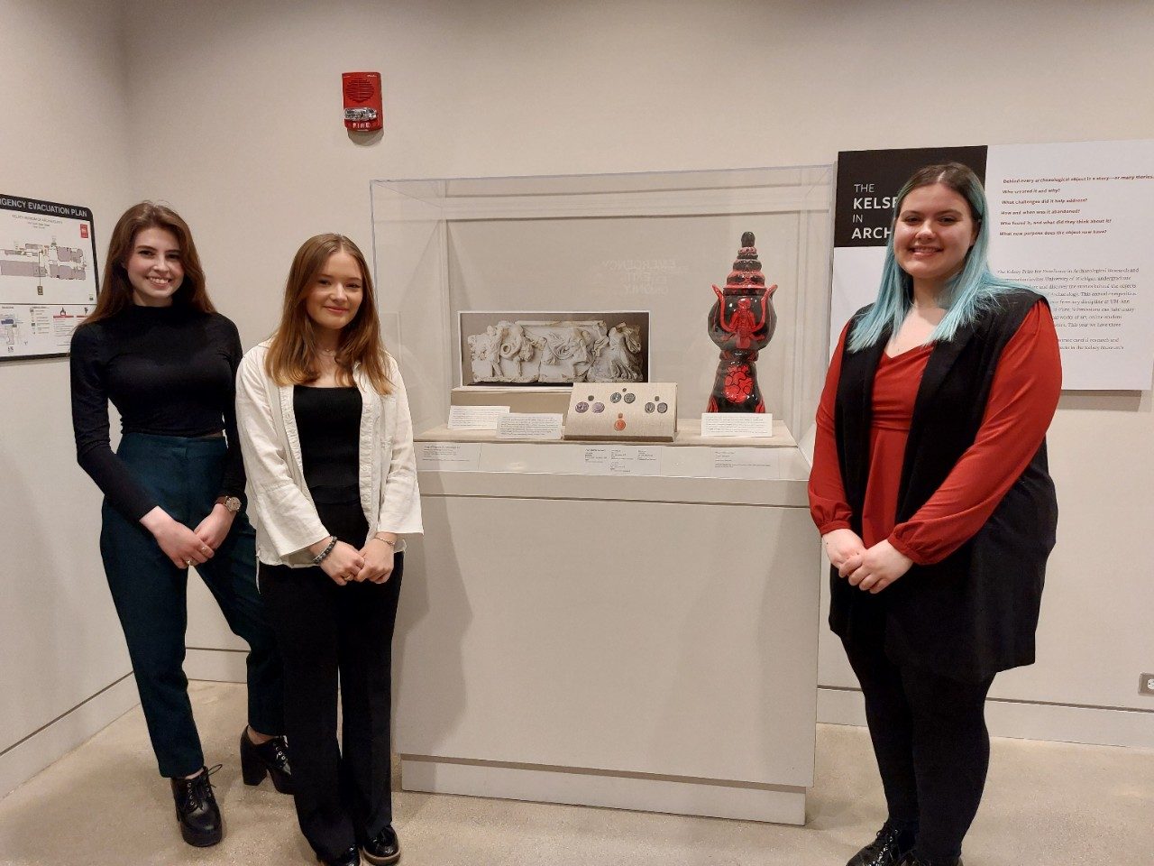 Three college-age women stand next to a museum display case containing photos of a carved marble sarcophagus lid and the obverse and reverse of four small oval objects, as well as a black and red pottery piece with painted images of a woman and anatomical heart. 