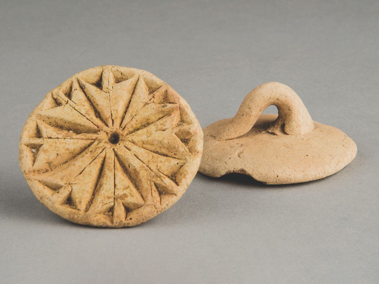 Two tan clay bread stamps rest side by side. One is flipped over, revealing its geometric pattern, while the other is facedown, showing a small handle or fingerhold.