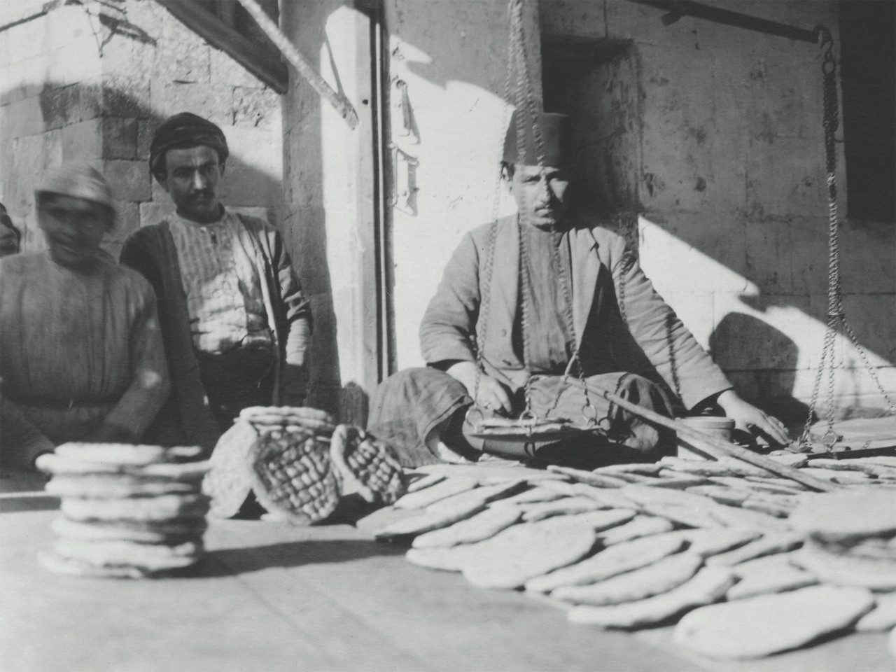 Three men stand behind a table covered with pieces of flat bread at a market. One, wearing a fez, weighs loaves on a scale. 