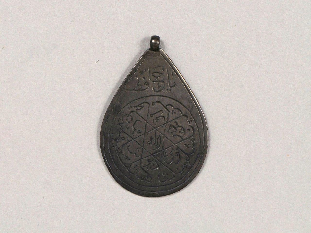 Teardrop-shaped amulet with a suspension loop at its top showing a circle containing Arabic inscriptions.
